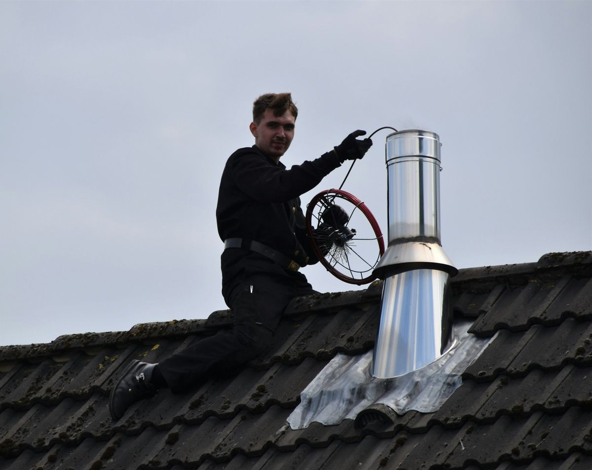 Een schoorsteenveger met een borstel op een dak, naast een stalen rookkanaal